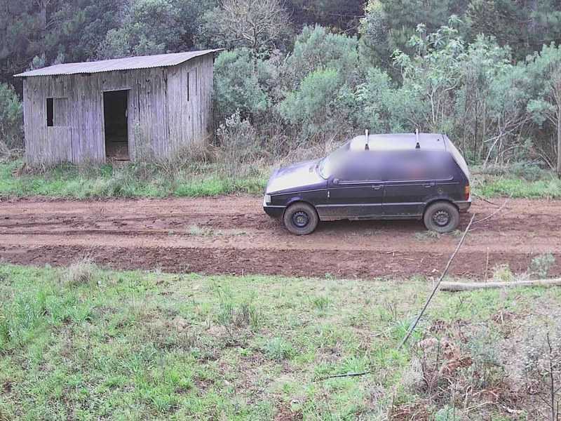 Carro passando pela estrada de terra em frente a um galpão de madeira